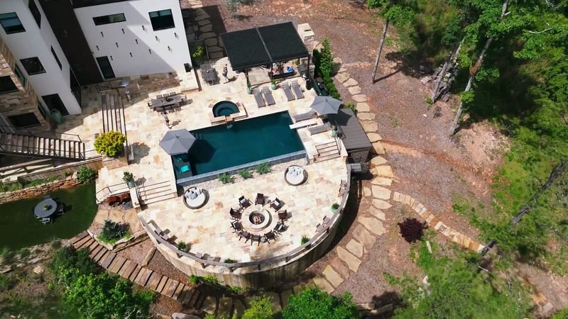 Aerial view of a stone patio with a rectangular pool, spa, fire pit, and pergola beside a modern home