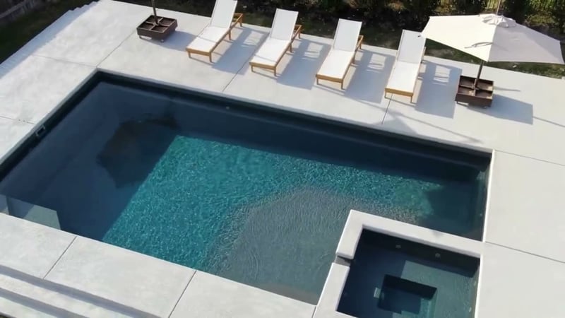 Aerial view of a modern rectangular fiberglass pool with an attached spa, concrete deck, and lounge chairs